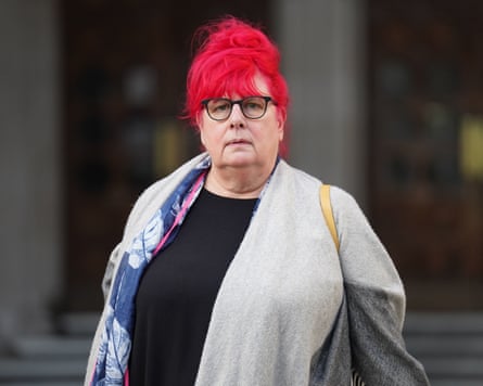 A middle-aged woman with glasses and red hair tied in a bun. She is looking directly at the camera and wearing a grey cardigan and a patterned scarf over a black top