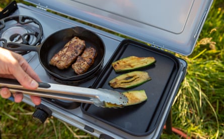 Someone turns courgettes on a Campingaz Camping Kitchen 2 Multi-Cook Plus Gas Stove while meat cooks in a pan on the unit