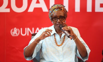 Picture of Winnie Byanyima sitting in front of red background with her hands raised to her face, talking into a microphone