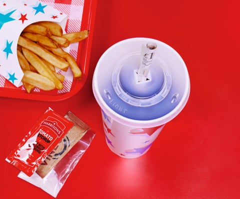 Packet of chips, ketchup sachets and drink holder with rolled up £10 note poking through hole in plastic lid, against red background