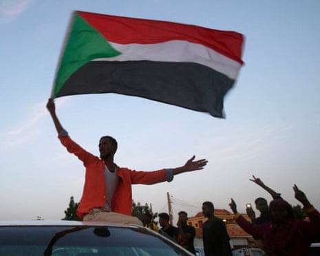 A man sitting on the roof of a car waves a Sudanese flag as other men give peace signs.
