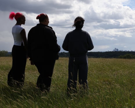 The three women stand with their backs to the camera on a wide expanse of grass overlooking the London skyline in the far distance