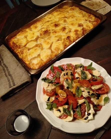 A colourful tomato salad on a white plate and a tray of lasagne on a wooden table