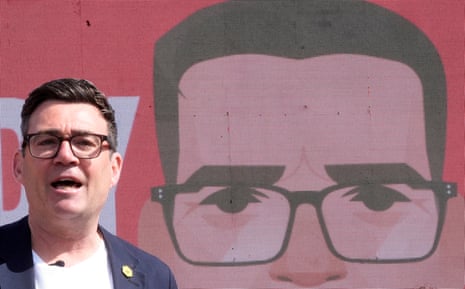 Andy Burnham speaks at a microphone in front of a large digital screen showing a cartoon face of himself