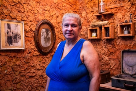 An older woman in a blue dress stands in a mud-walled room with old photos and a crucifix hanging on it