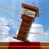 A tilted Turning Point USA podium pulled by ropes from both sides against a heavenly blue sky