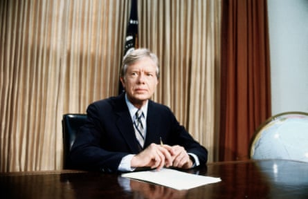 A man sits at a desk in front of a flag