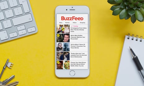 An iPhone showing the BuzzFeed website rests on a yellow background table with a keyboard, keys, notepad and plant