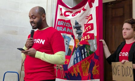 Shaban Ali stands reading from a phone into a microphone with a protest banner being held behind him