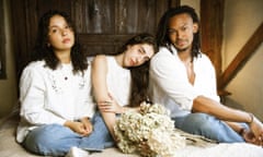 Cinya Khan, Maya Rae and Judah Mayowa, dressed in jeans and white shirts, sitting on a bed with some dried flowers