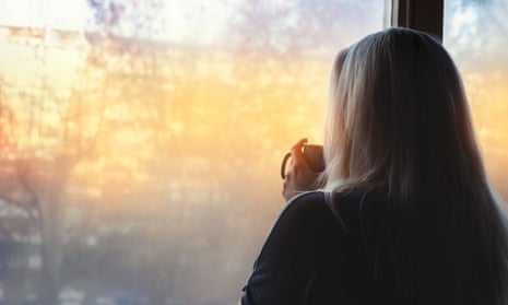 Posed by model Blonde woman standing by the window, with coffee cup in hands, looking out into the morning light