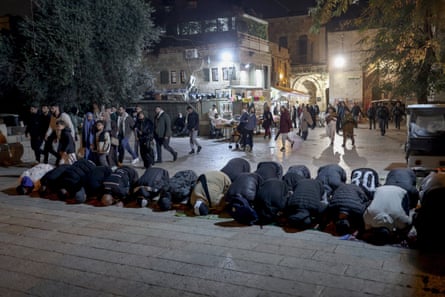 Palestinian worshippers kneel at the steps to the mosque, some on small prayer mats. Families and other people are moving behind them and there are market stalls in the background by tall stone walls and buildings.