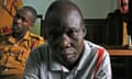 A middle-aged African man stares at the camera as police sit behind him in a courtroom