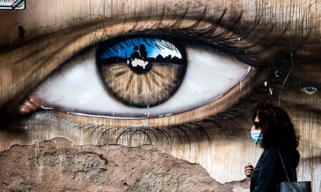A person wearing a face mask walks in front of a mural of a large eye during the Covid pandemic in Italy, Rome.