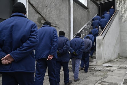 A row of men in blue jackets and black hats walk with backs to camera towards and up a staircase and through an open door to a building