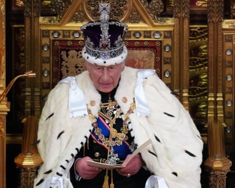 King Charles III in ceremonial robes and crown reads from papers while seated on an ornate throne