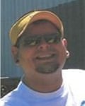 A blurry picture of a young-ish white man wearing sunglasses and a yellow visor, with a brown goatee and black stud earrings in both ears, smiling in a white T-shirt.