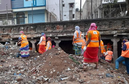About 20 Indian women and men standing on a mound of soil and rubbish in a open space below ground surrounded by a colonnade