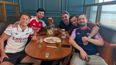 Five men sitting at a table with beer, two of them wearing Arsenal shirts