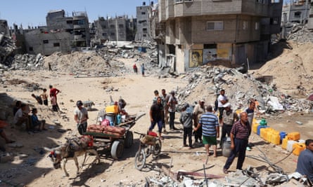 Displaced Palestinians collect water in containers, surrounded by rubble and destroyed buildings, with a donkey pulling a loaded cart
