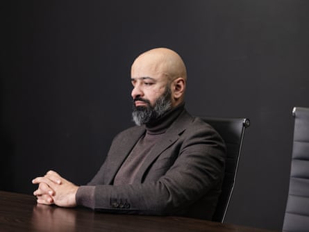 A solemn-looking man pinch a beard and shaved caput successful a general overgarment and polo cervix jumper sits astatine a table