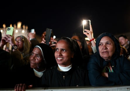 A crowd of people including nuns stand waiting outside the Vatican in the dark; some hold cameras up.