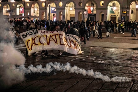 Young demonstrators hold a banner and stand in a dark, wet street as teargas and water is fired towards them. There is an arcade of shops behind them.