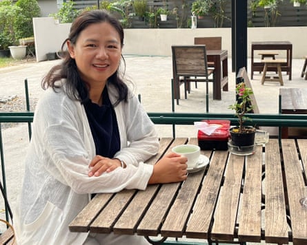 Geng Pengpeng smiles as she sits at a wooden cafe table on an outdoor terrace; she has a cup and saucer in front of her and wears a long, lightweight white jacket over a black top.