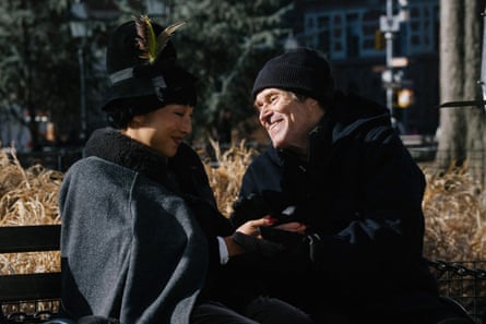Lee and Dafoe, both dressed in black coats and hats, sit holding hands on a park bench