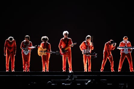 David Byrne and his group wearing orange clothes stand in front of a black background playing their instruments