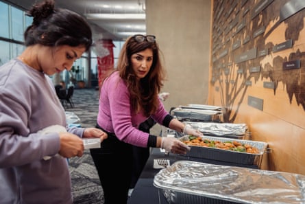 a woman put down a tray of food on a table as another woman stands by reading a paper