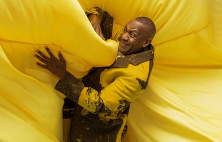 Lenny Henry in a yellow and brown jacket, grappling with a billowing background of yellow fabric