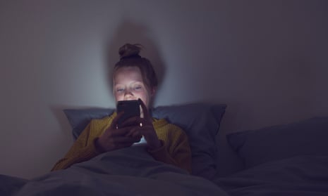 A young teenager is sitting in her bed in the dark with only the light of her phone shining on her face.