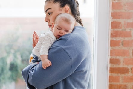 A sad looking young woman standing at a window with a sleeping baby lying against her shoulder.
