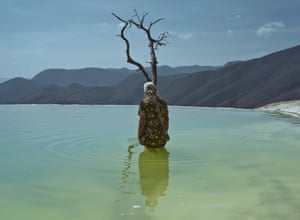 An elderly woman stands thigh-deep in still blue-green water, by a dead tree