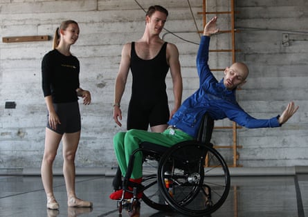 Two dancers stand next to Mark Brew, who has his arms uplifted while he uses a wheelchair.