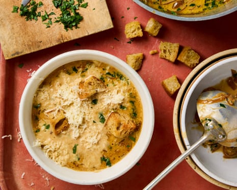 A bowl of onion and parsley soup on a reddish tray, alongside some scattered croutons, parsley on a chopping board and a large pot of soup.