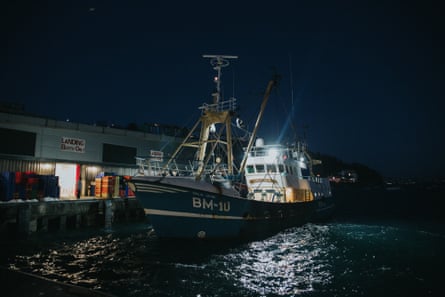 A large fishing boat tied up at a quay by a warehouse at night