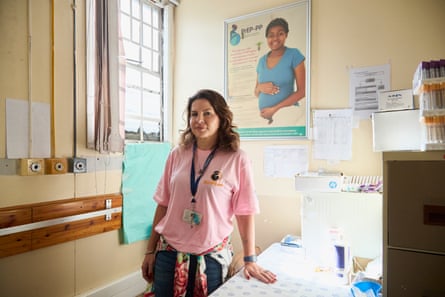 A doctor in a pink T-shirt stands facing the viewer in a consultation room, medical supplies piled on a filing cabinet to one side of her and information sheets pinned to the back wall.