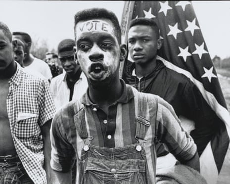 A young civil rights marcher with "VOTE" painted on his forehead, a person carrying an American flag behind him