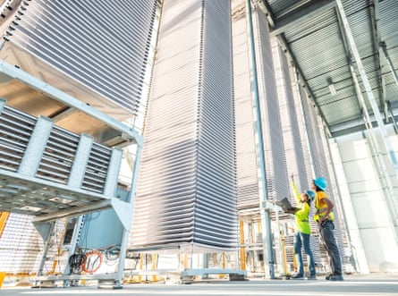 Two people in hard hats and safety vests stand beside seemingly sky-high towers of trays inside a metal barn-like, lighted facility.