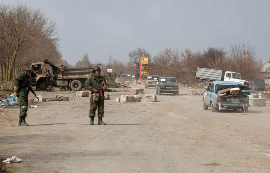 Two pro-Russian troops stand guard at a checkpoint. Three cars drive into the distance - nearest car has boot overloaded with luggage