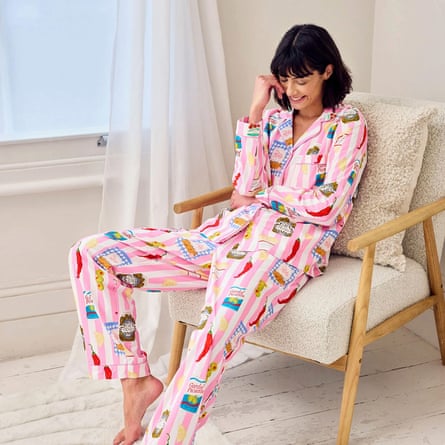 A model is smiling happily in a soft chair by a window wearing pink and white striped Chelsea Peers pyjamas with kitsch print