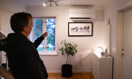 A man stands in a room in his house pointing to a white heat pump fixed to the wall above a picture