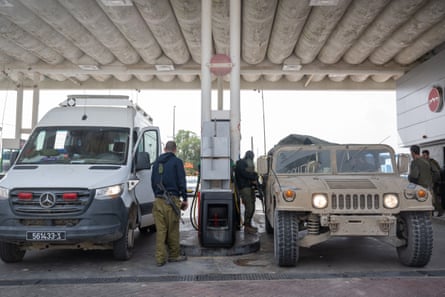 Under the metal canopy of a gas station, two a beige open-roofed Hummer, which looks like a jeep, and a white Mercedes van both fill up their vehicles.