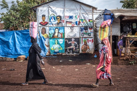 Two women carrying sacks on their heads walk past a refugee camp tent decorated with portraits and aid organisation logos