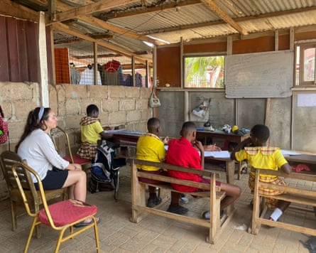 A young woman sitting behind young children on benches in a very simple classroom