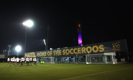 Jugadores en la cancha iluminada, con un cartel gigante que dice Home of the Socceroos en el fondo