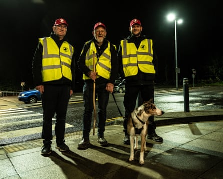 Three smiling men in hi-vis jackets with a dog on a street corner at night
