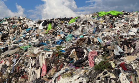 A large landfill displays a mountain of discarded textiles and mixed waste, with vibrant clothing colors contrasting against a blue sky and white clouds